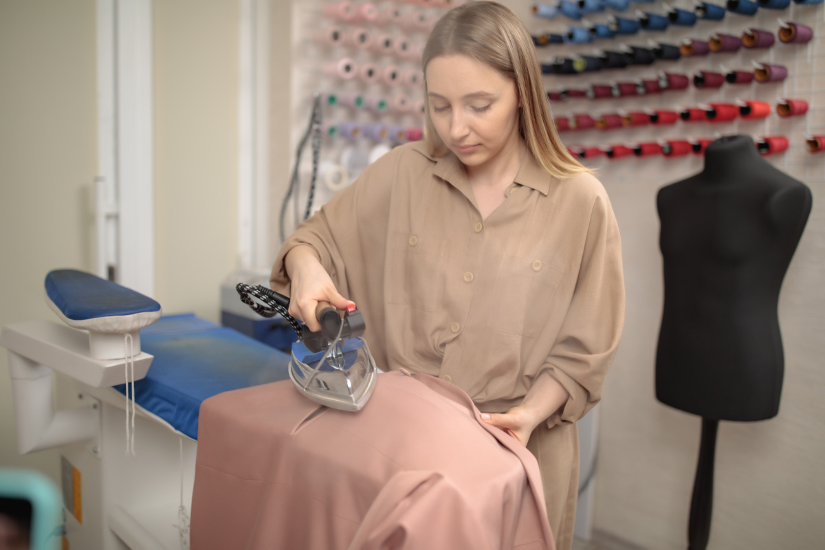 A Seamstress Girl Is Standing In A Studio Near An Ironing Board And Ironing Clothes. Workplace. Creation Of Fashion Clothes In The Studio A Seamstress Girl Is Standing In A Studio Near An Ironing Board And Ironing Clothes. Workplace. Creation Of Fashion Clothes In The Studio